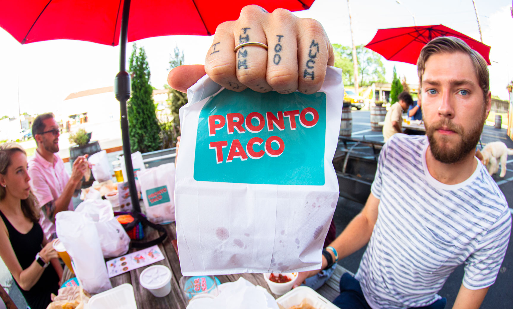 Fish eye lens photo of tattooed hand holding Pronto Taco bag in foreground with people eating outside in background